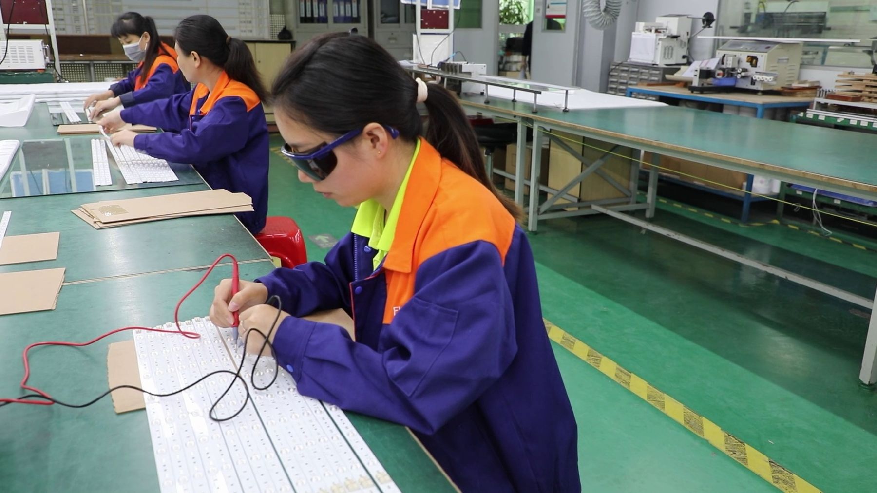 A worker in safety goggles testing an LED board with meter probes at a factory bench